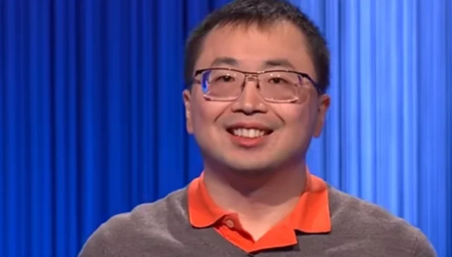 Smiling man with glasses in a gray sweater and orange polo, standing against blue backdrop curtains on a stage.