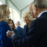 Businesspeople greeting with a handshake at a formal event; woman in blue jacket on the left, man in a dark suit on the right.