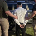 Man in a white shirt with hands cuffed being led by two officers beside a SUV, one wearing POLICE Homeland Security Investigations vest and the other a U.S. Marshal vest.