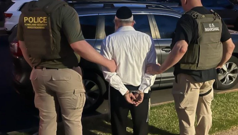 Man in a white shirt with hands cuffed being led by two officers beside a SUV, one wearing POLICE Homeland Security Investigations vest and the other a U.S. Marshal vest.