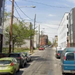Residential street lined with parked cars on both sides, modern multi-story buildings, and overhead power lines under a clear blue sky; a person stands near a sidewalk on the right.