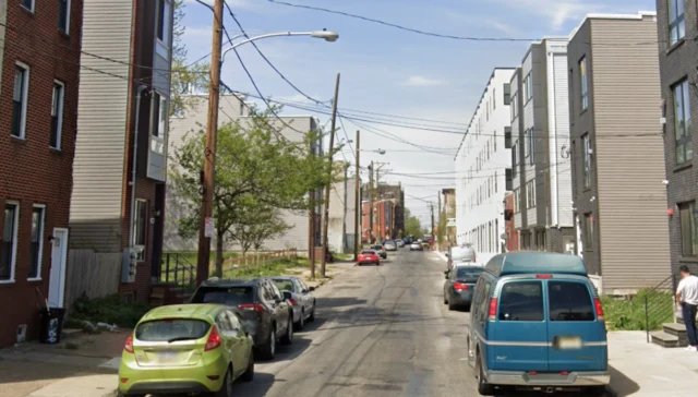 Residential street lined with parked cars on both sides, modern multi-story buildings, and overhead power lines under a clear blue sky; a person stands near a sidewalk on the right.
