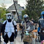 Four people in costume posing outdoors at a cafe, including Mandalorian armor and Baby Yoda prop, with string lights overhead.