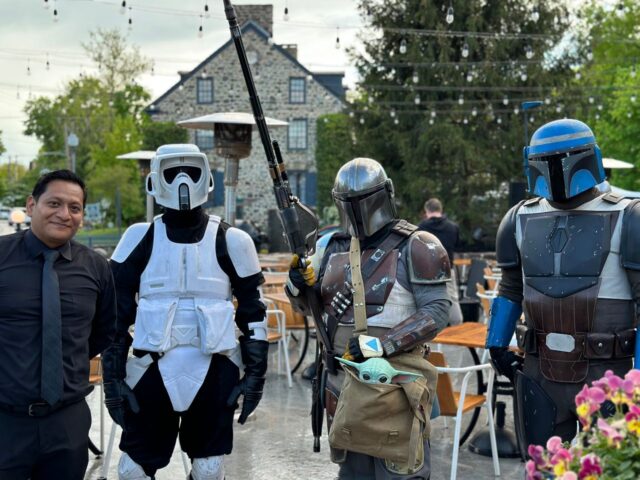 Four people in costume posing outdoors at a cafe, including mandalorian armor and baby yoda prop, with string lights overhead.