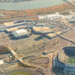 Aerial view of a large sports complex with a circular stadium, surrounding buildings, parking lots, and a river in the distance.