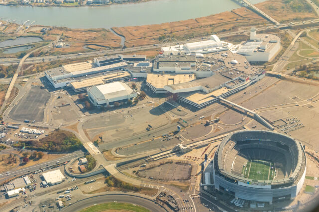 Aerial view of a large sports complex with a circular stadium, surrounding buildings, parking lots, and a river in the distance.
