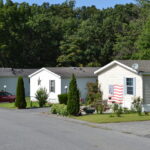 Row of white mobile homes with manicured yards and an American flag décor in the foreground, backed by dense green trees.