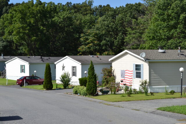 Row of white mobile homes with manicured yards and an american flag décor in the foreground, backed by dense green trees.