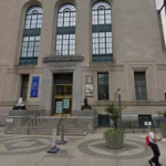 Facade of The Newark Museum of Art with tall arched windows, banners, and stone steps on a city sidewalk; a person walks by and patterned pavement circles the entrance.