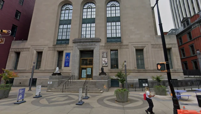 Facade of the newark museum of art with tall arched windows, banners, and stone steps on a city sidewalk; a person walks by and patterned pavement circles the entrance.