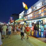 Bustling nighttime boardwalk scene with souvenir shop selling colorful t‑shirts, signs, and crowds of shoppers.