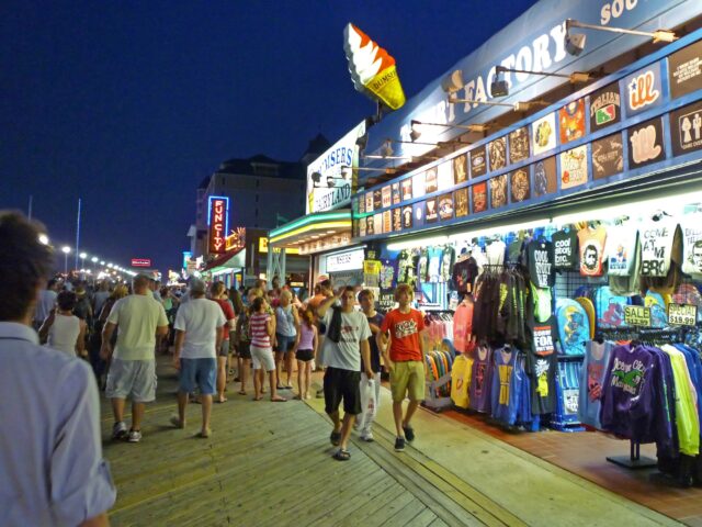 Bustling nighttime boardwalk scene with souvenir shop selling colorful t‑shirts, signs, and crowds of shoppers.