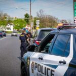 Two police officers talk beside a marked police SUV on a busy suburban street with a green highway sign in the background.