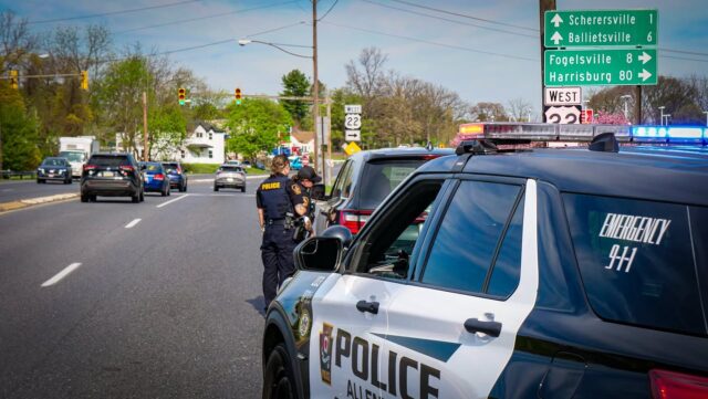 Two police officers talk beside a marked police suv on a busy suburban street with a green highway sign in the background.