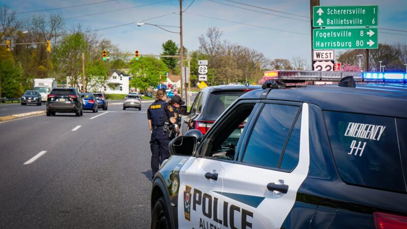 Two police officers talk beside a marked police suv on a busy suburban street with a green highway sign in the background.