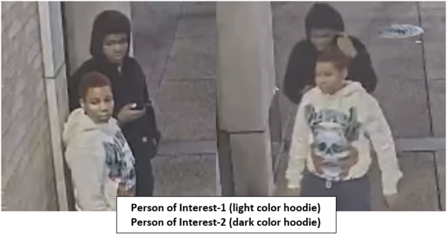 Two teens near a doorway; one in a white hoodie faces the camera, the other in a black hoodie looks at a phone.
