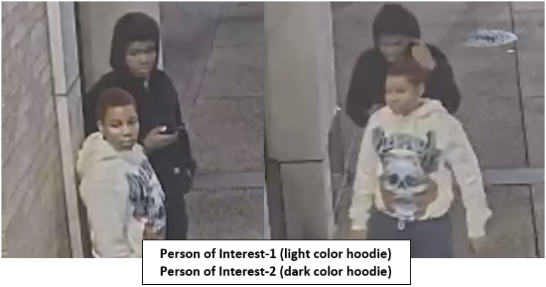 Two teens near a doorway; one in a white hoodie faces the camera, the other in a black hoodie looks at a phone.