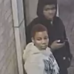 Two teens near a doorway; one in a white hoodie faces the camera, the other in a black hoodie looks at a phone.