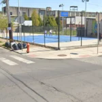 Urban street corner with a blue fenced basketball court and colorful mural walls; a few people stand inside the court area.