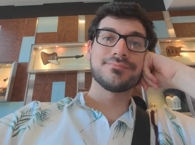 Young man with glasses rests his chin on his hand in a guitar showroom with display cases behind him.