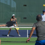 Four adults play pickleball on an outdoor court, two players on each side near the net with paddles ready and a ball in play.