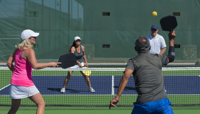 Four adults play pickleball on an outdoor court, two players on each side near the net with paddles ready and a ball in play.