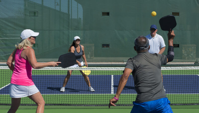 Four adults play pickleball on an outdoor court, two players on each side near the net with paddles ready and a ball in play.