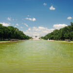Wide view of the Reflecting Pool with green trees on both sides and the Lincoln Memorial in the distance under a blue sky.