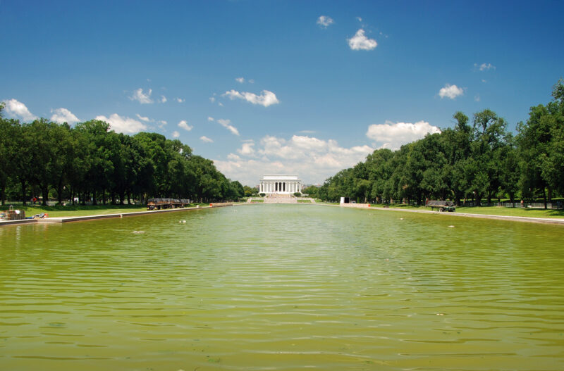 Wide view of the reflecting pool with green trees on both sides and the lincoln memorial in the distance under a blue sky.