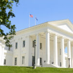 White neoclassical building with tall columns, flagpoles, and blue sky; green lawn in foreground; tree branches frame the left side.