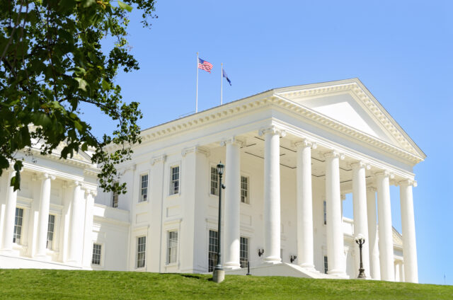White neoclassical building with tall columns, flagpoles, and blue sky; green lawn in foreground; tree branches frame the left side.
