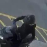 Motorcyclist in dark clothing riding a motorcycle past yellow parking-lot lines, viewed from above/side angle in motion.