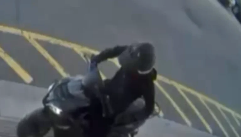 Motorcyclist in dark clothing riding a motorcycle past yellow parking-lot lines, viewed from above/side angle in motion.