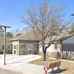 Public, single-story building with wheelchair ramp and benches outside, under a blue sky with a bare-tree foreground and a partial flag in the corner.