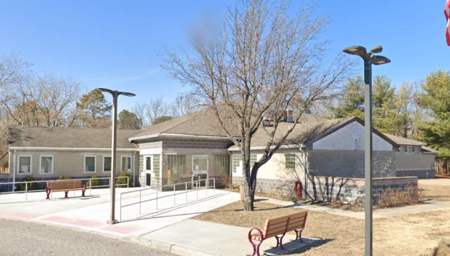 Public, single-story building with wheelchair ramp and benches outside, under a blue sky with a bare-tree foreground and a partial flag in the corner.