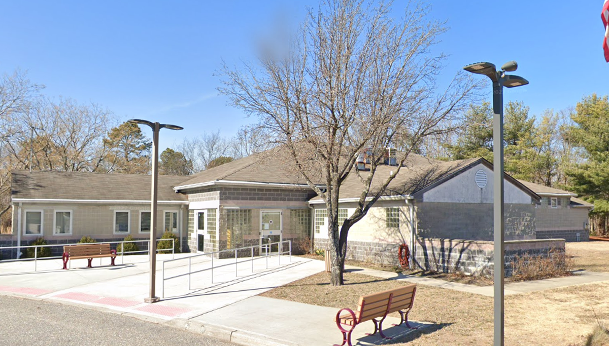 Public, single-story building with wheelchair ramp and benches outside, under a blue sky with a bare-tree foreground and a partial flag in the corner.