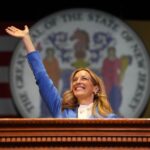 Female politician at a podium, hand raised in a-wave, with a state seal backdrop behind her in a formal setting.