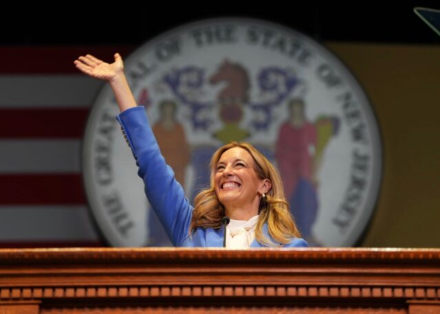 Female politician at a podium, hand raised in a-wave, with a state seal backdrop behind her in a formal setting.