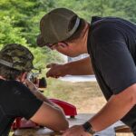 Two men at an outdoor shooting range; one guides the other as they aim a rifle at a bench.