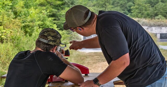 Two men at an outdoor shooting range; one guides the other as they aim a rifle at a bench.