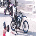 Person posing for a photo beside a black dirt bike on a city sidewalk with others and a stroller in the background.