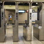 Row of stainless steel subway turnstiles in a station, with pipes overhead and a map visible in the background.