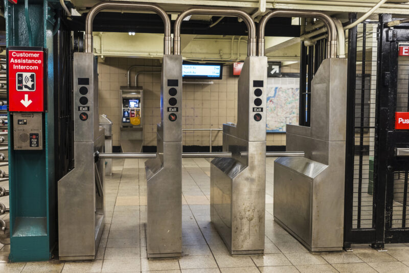 Row of stainless steel subway turnstiles in a station, with pipes overhead and a map visible in the background.