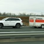 White SUV towing a U-Haul trailer on a highway with guardrails and dry roadside vegetation in the background.
