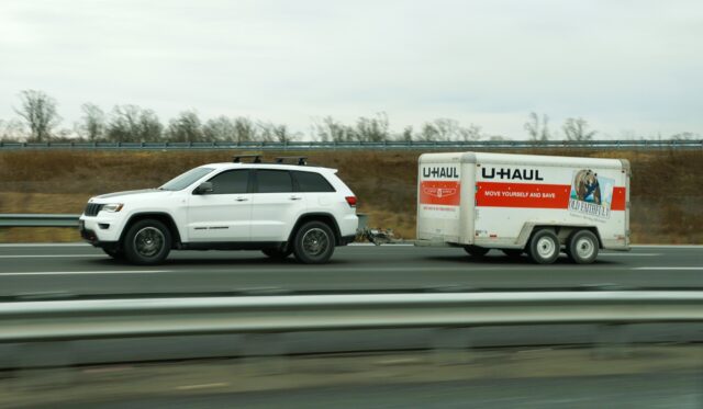 White suv towing a u-haul trailer on a highway with guardrails and dry roadside vegetation in the background.