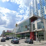 Urban street with a modern glass office building, several cars, and pedestrians under a blue sky with clouds.