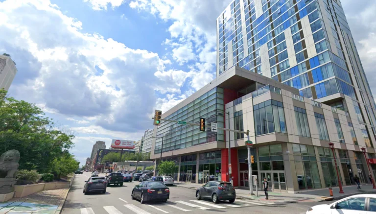 Urban street with a modern glass office building, several cars, and pedestrians under a blue sky with clouds.
