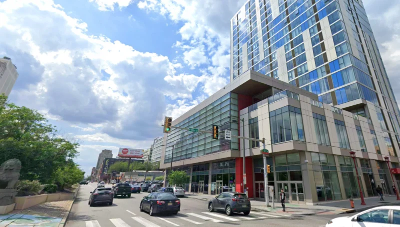 Urban street with a modern glass office building, several cars, and pedestrians under a blue sky with clouds.