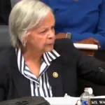 Female official in a dark blazer and striped blouse speaks into a microphone at a conference table; nameplate partially visible.