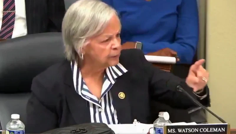 Female official in a dark blazer and striped blouse speaks into a microphone at a conference table; nameplate partially visible.